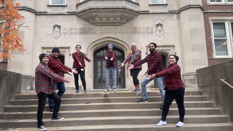 Members of the lab standing on stairs in front of the medical laboratories making fun poses and smiling.